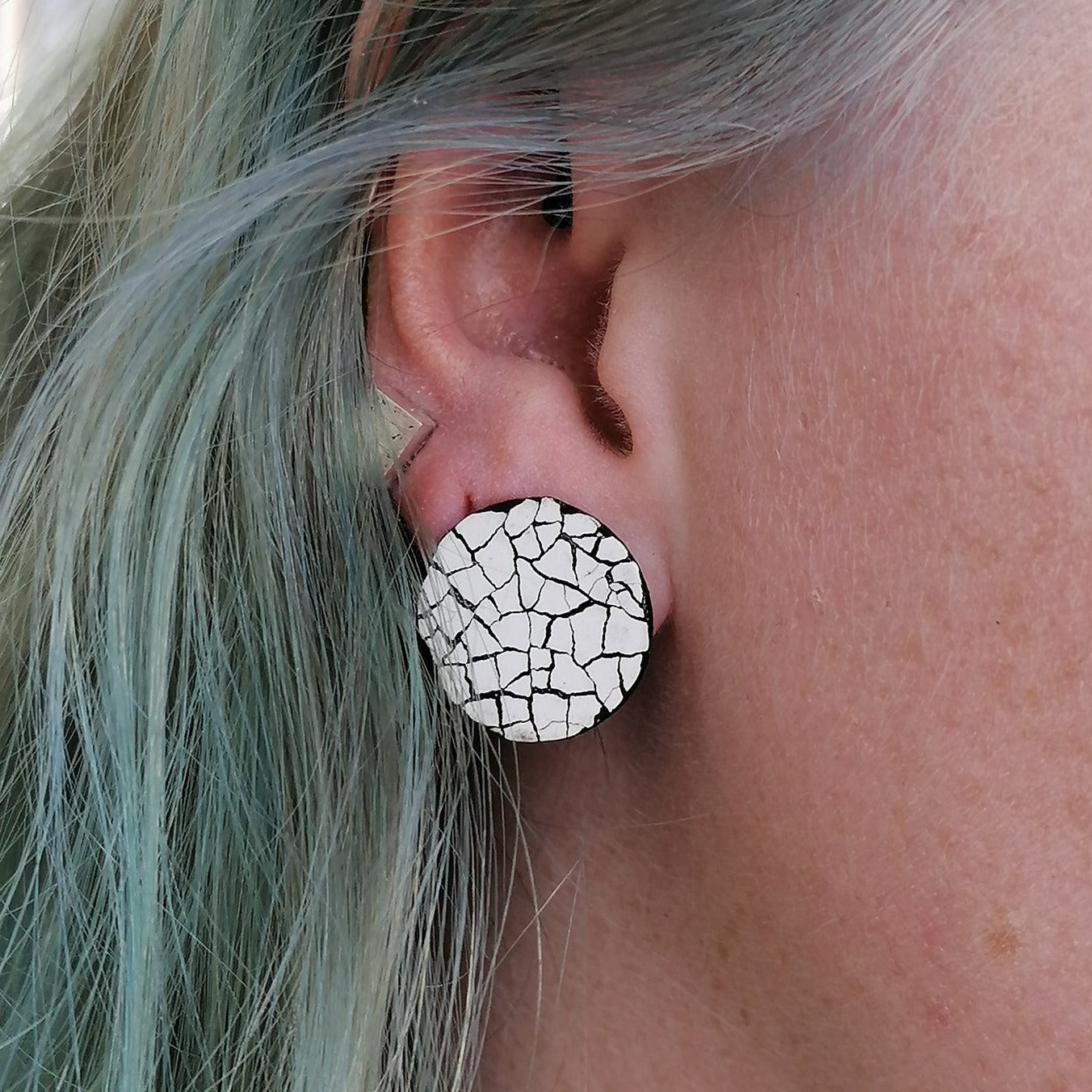 Image shows a single round stud modern mosaic earring made using the Rankaku technique using real eggshell, white on black. The earring is being worn by a blue haired model.