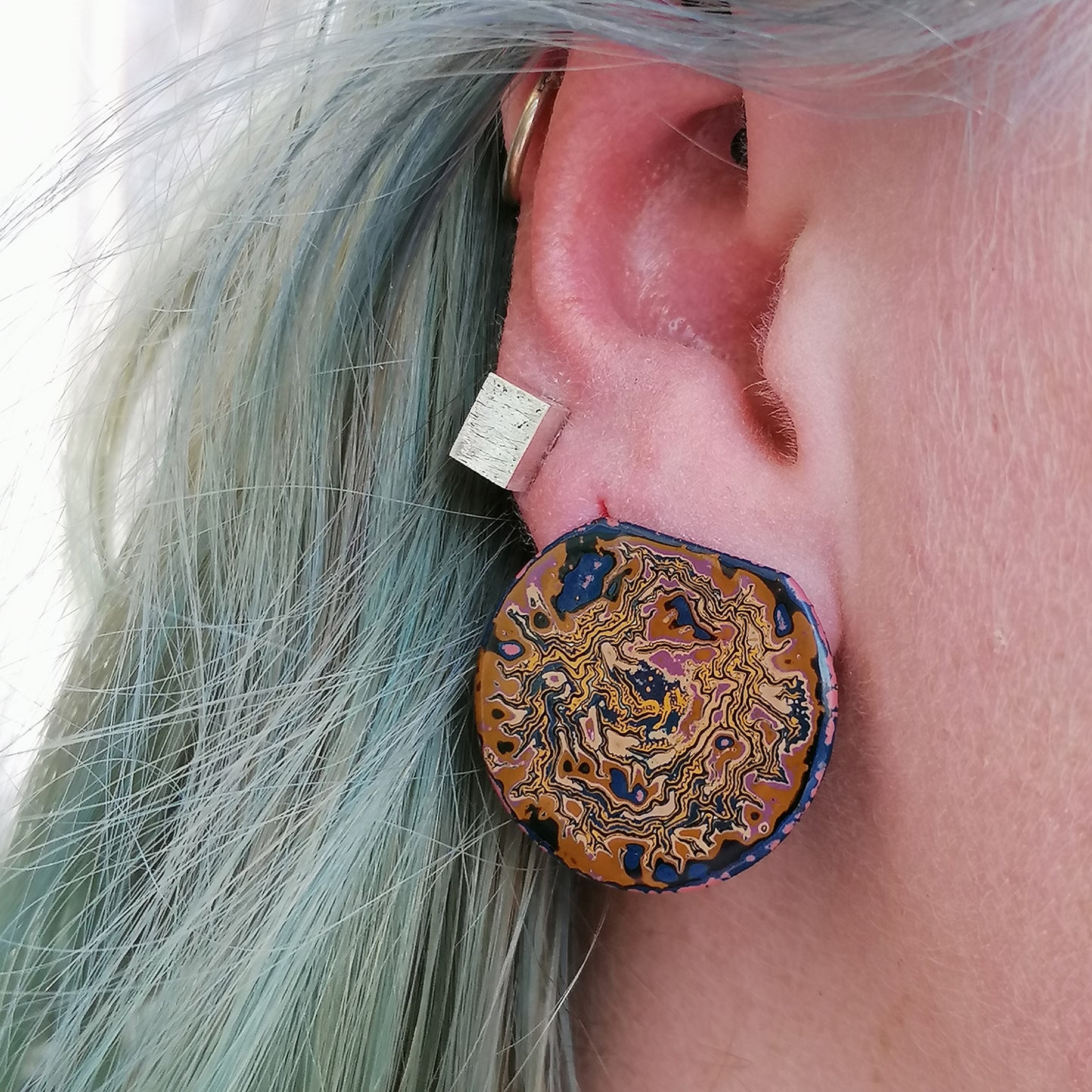 image shows a single round stud earring with the top portion cut off. Earring shows a wrinkled pattern with yellow deep blue and beige. The earring is being worn by a blue haired model.