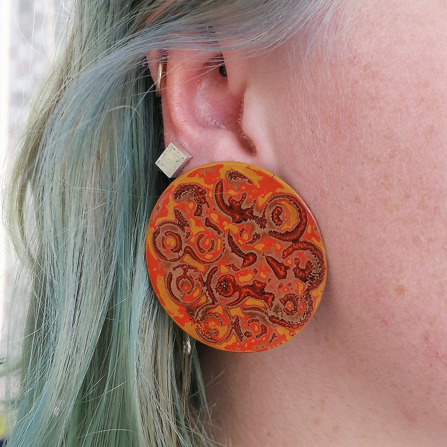 Image shows a statement earring being modeled by a model with blue hair. Pattern consists of circles in orange, brown, yellow and a pale pink.