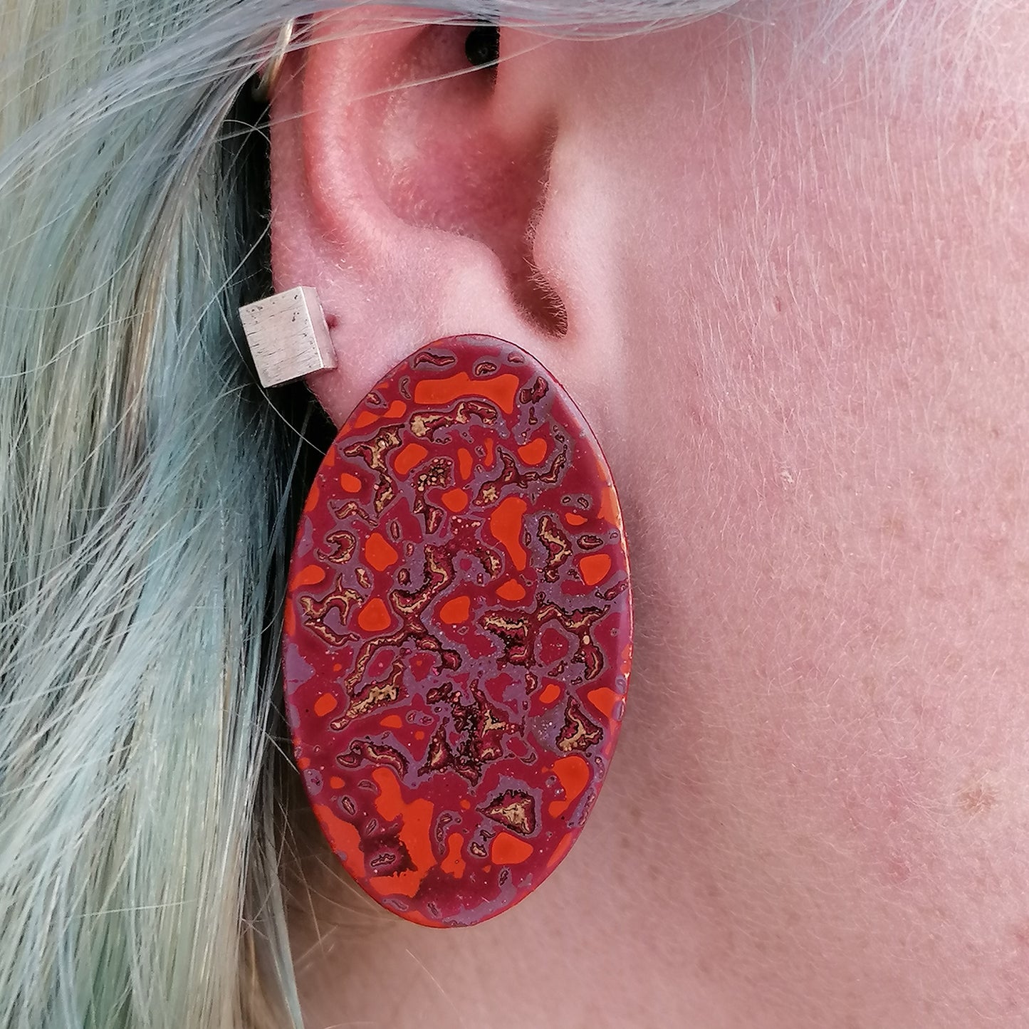Image shows a single long oval earring with a pattern in pink, purple, orange and white. The earring is being modeled by a blue haired model