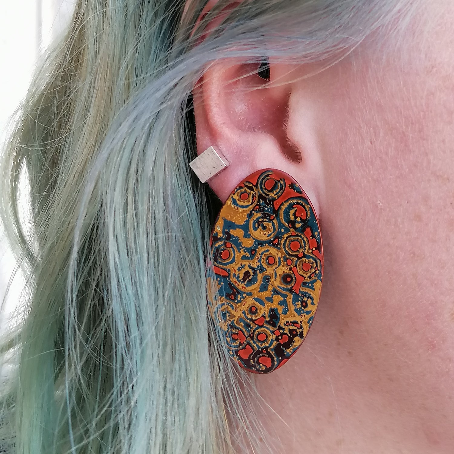 Image shows a long oval statement earring being worn by a blue haired model. The pattern consists of rings in yellow, blue and orange