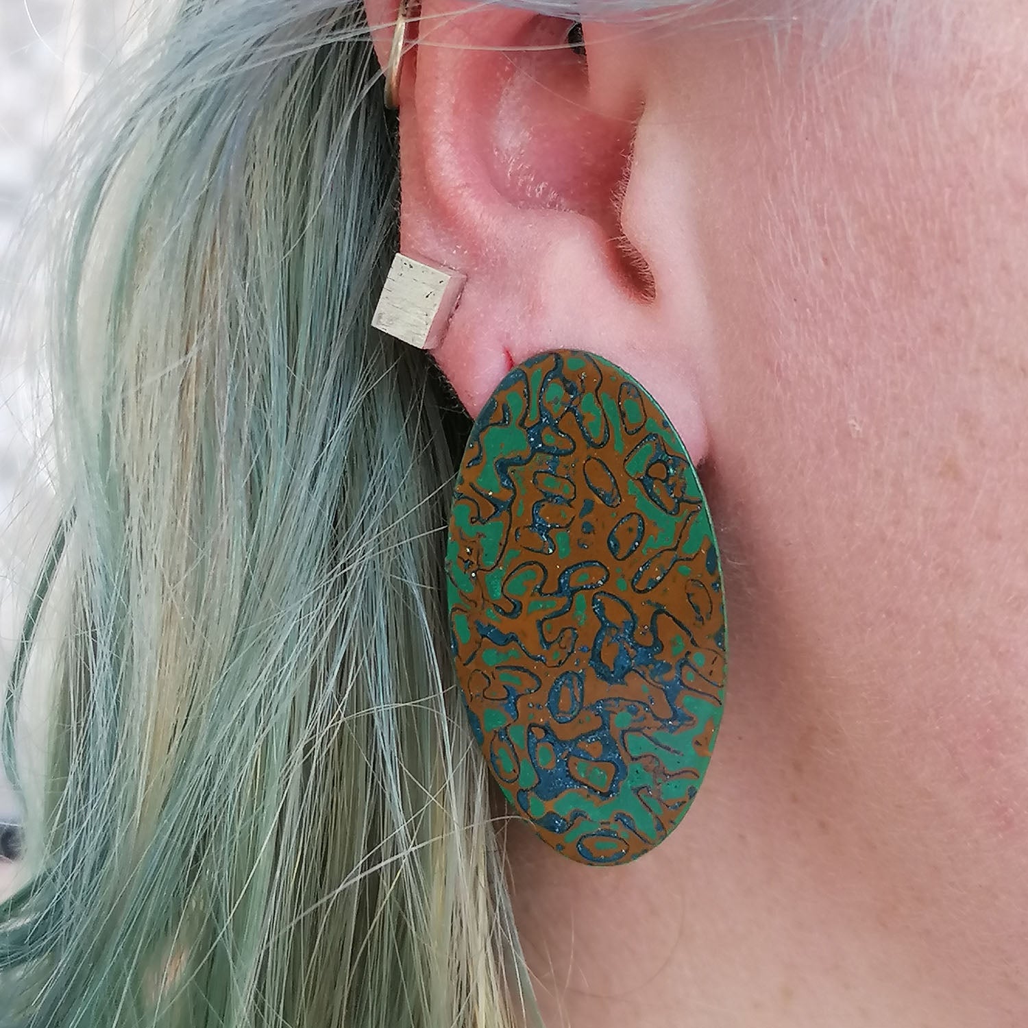 Image shows a single long sleek and shiny statement oval earring being worn by a blue haired model. Abstract pattern in ochre yellow, green and blue