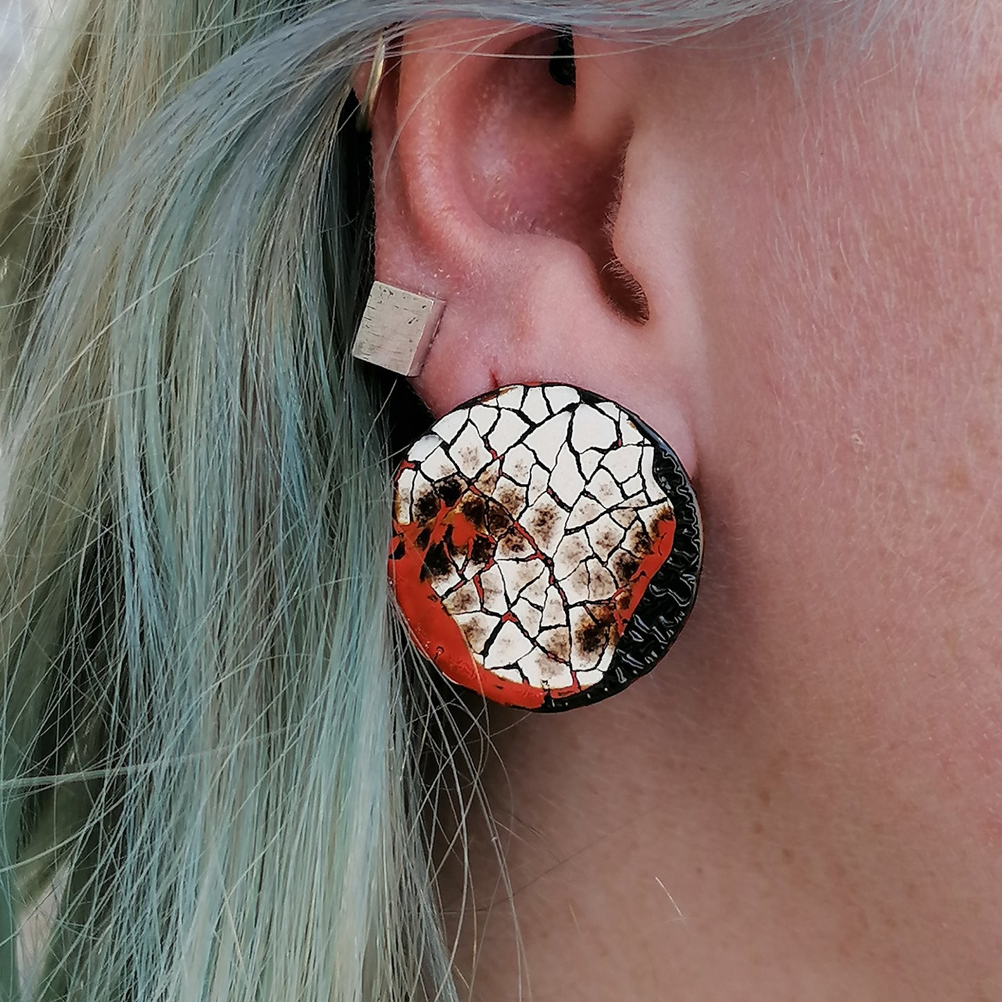 Images shows a single round stud earring being modeled by a blued haired model.. Modern mosaic earrings made out of real eggshell and black lacquer with an area of raised black wrinkled texture and patches of red.