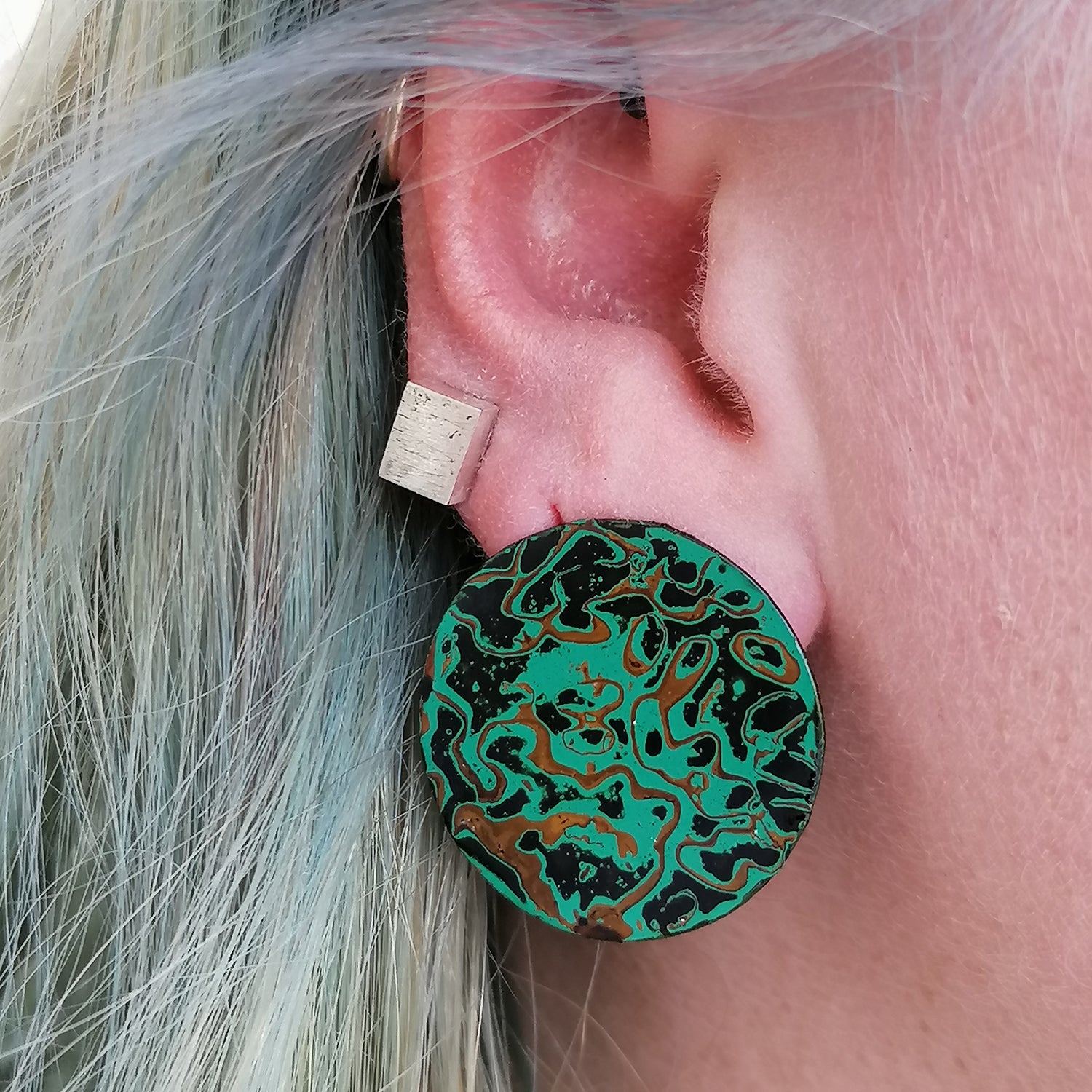 Image shows a single circle stud earring being worn by a blue haired model. An abtract pattern in black, green and ochre yellow