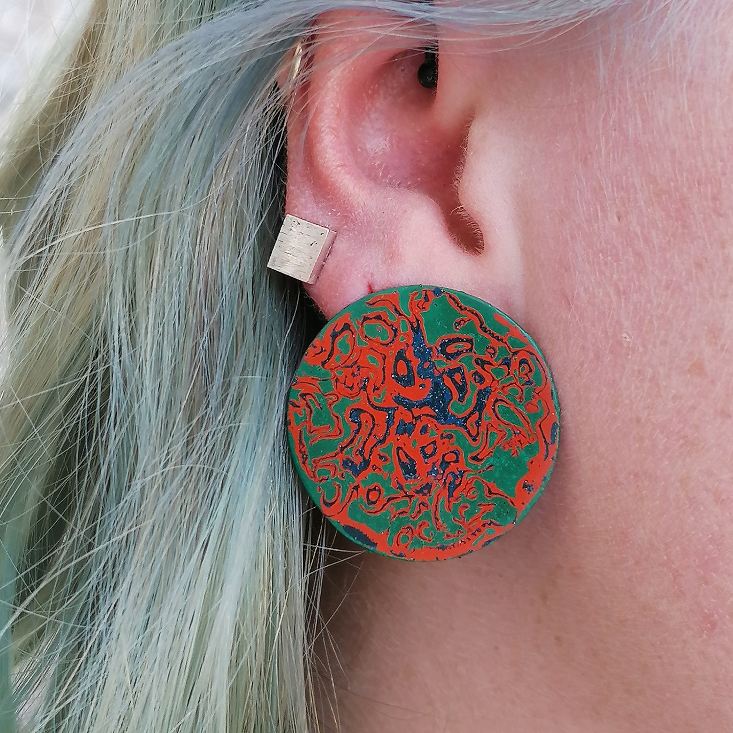 image shows a pair of circle stud earrings in green, orange and blue, being worn by a blue haired model.