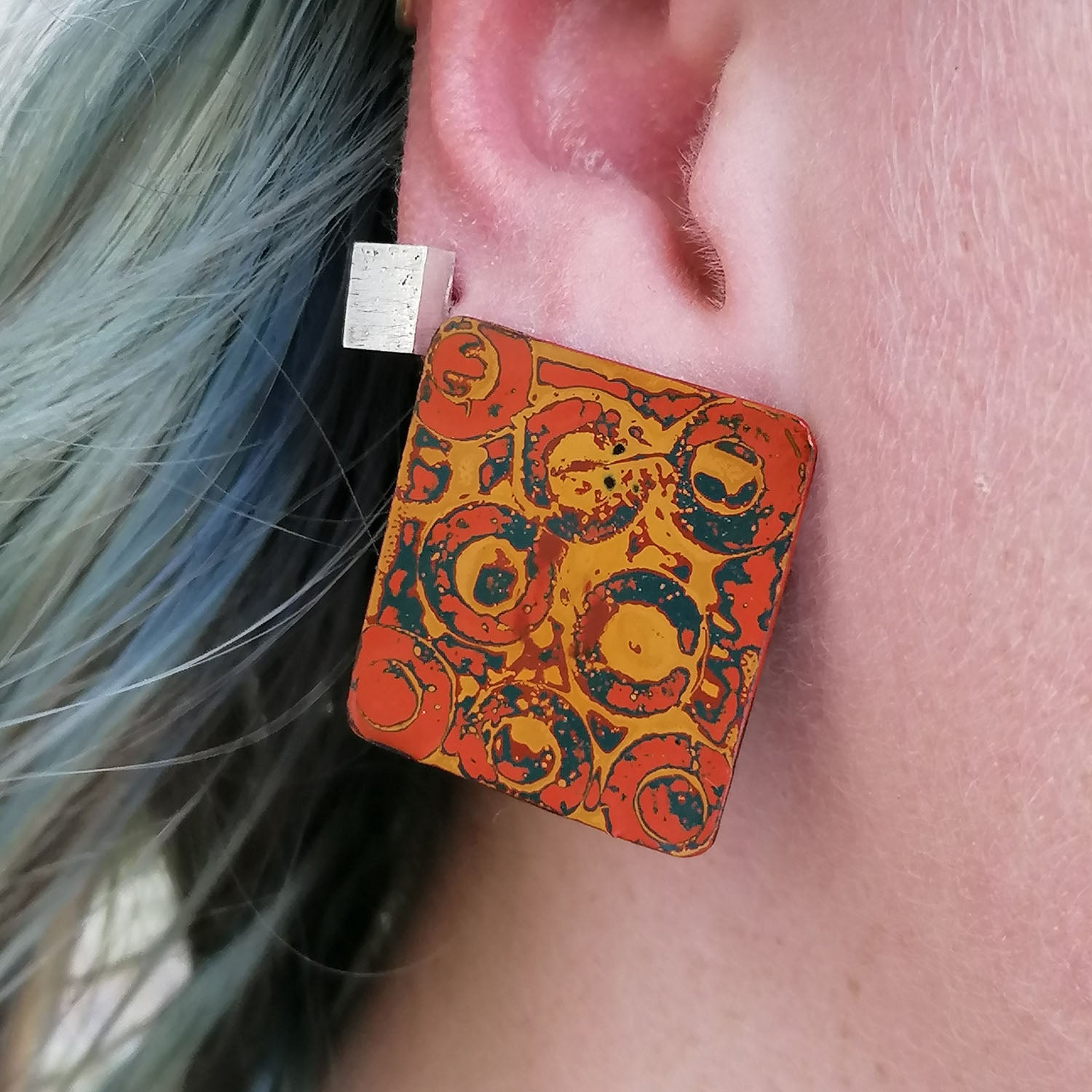 Image shows a pair of soft square earrings being worn by a blue haired model with an abstract pattern in circular pattern in orange, yellow and blue