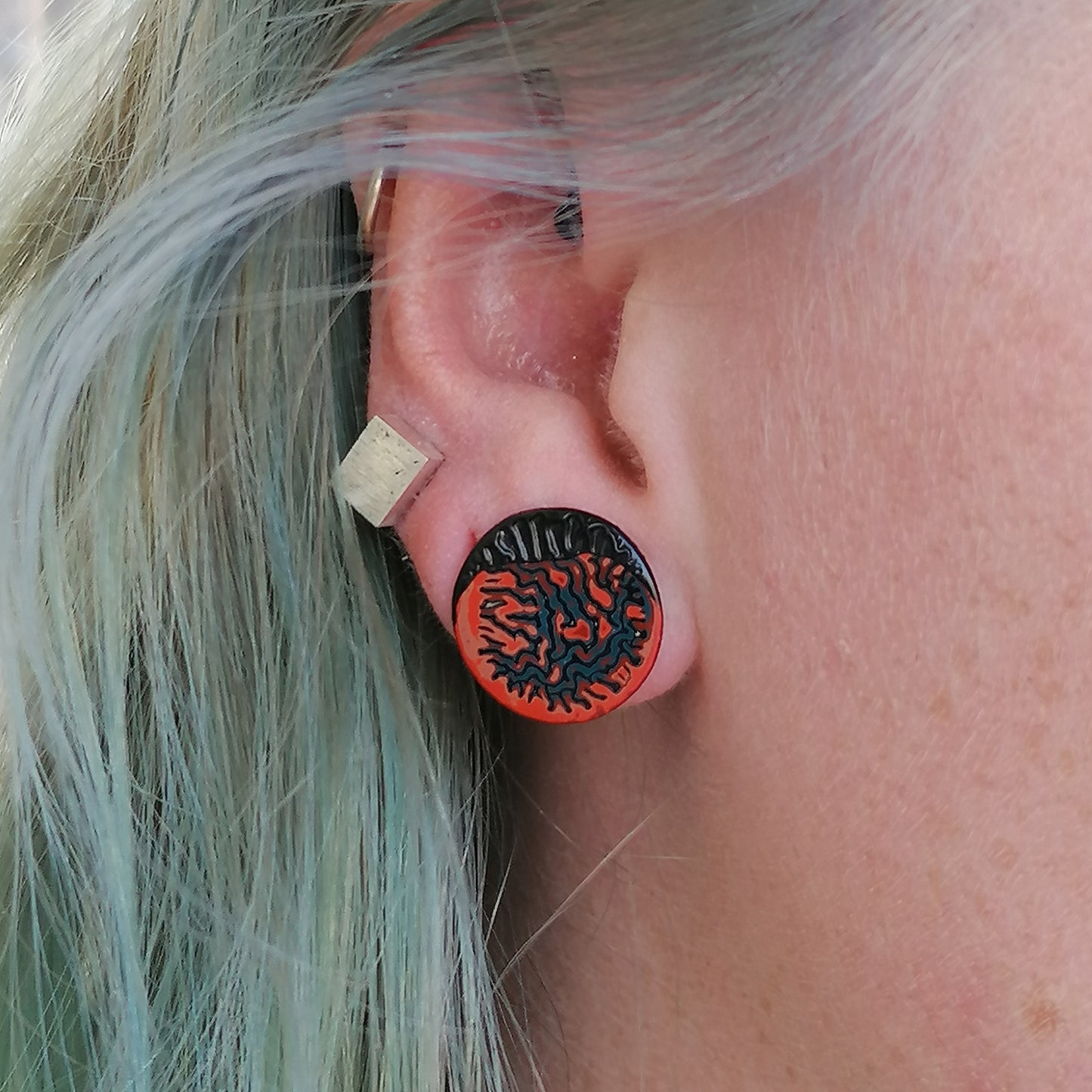 Image shows a single circle stud earring being worn by a blue haired model. With a wrinkled pattern in orange, blue and red and a raised black winkle textured rim.