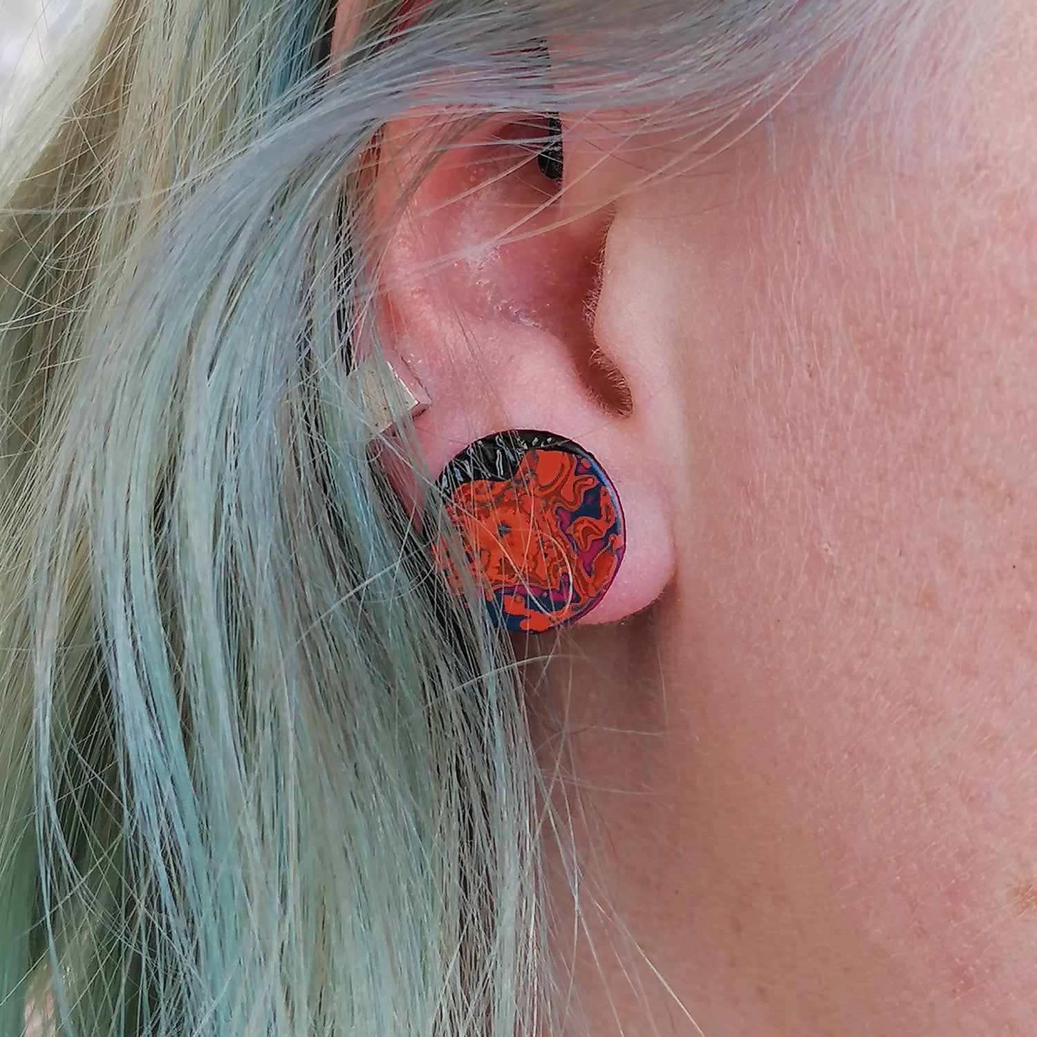 Image shows a single small circle stud earring being worn by a blue haired model. An abstract pattern in pink, orange and blue.