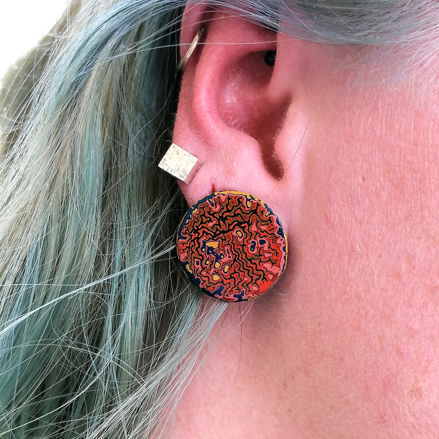 Single circle stud earring being worn by a blue haired model. Wrinkled pattern in orange, pink and blue.